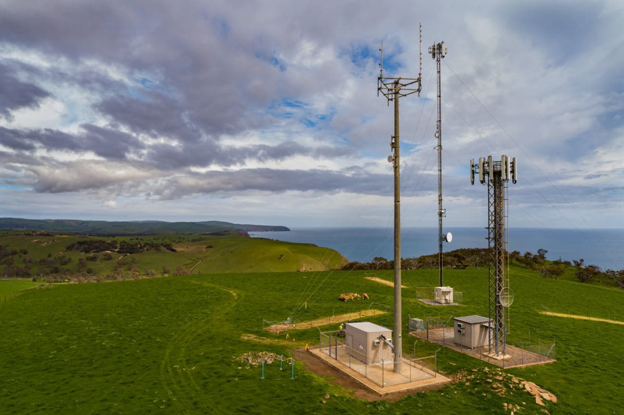 Three telecommunication towers on a flat stretch of grass with hills, ocean, and sky in the background.